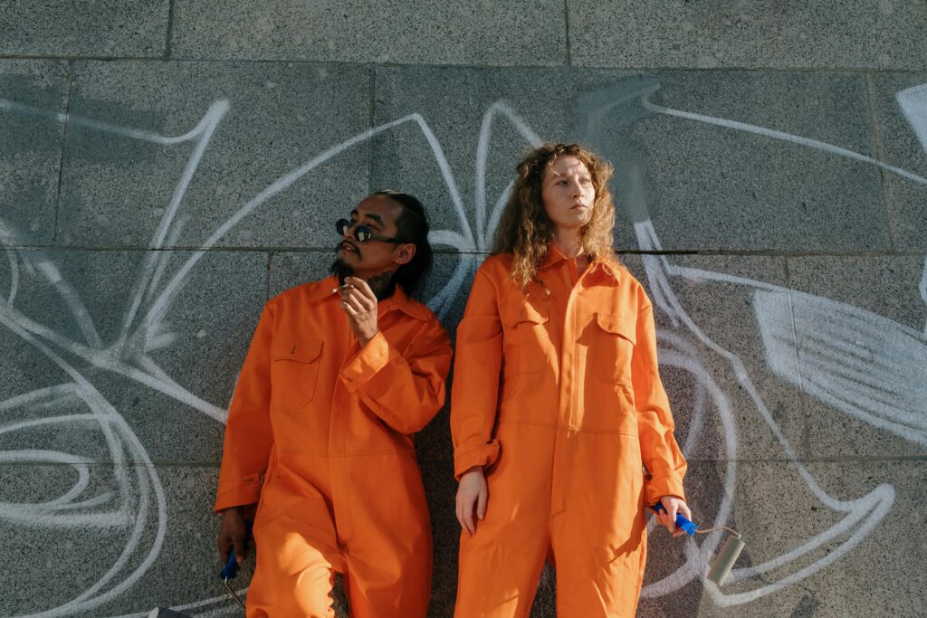 Two community workers in orange uniforms standing against a graffiti wall, holding paint rollers outdoors.
